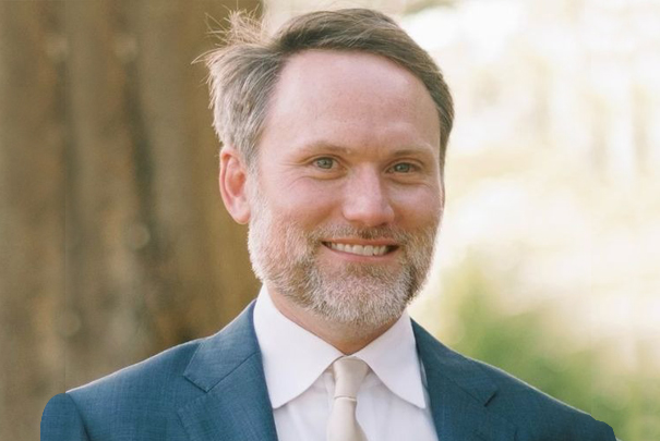 Professional headshot of smiling man in blue blazer and light shirt outdoors.