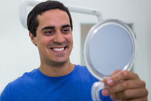 Man in blue shirt smiling while holding handheld mirror in dental office setting.