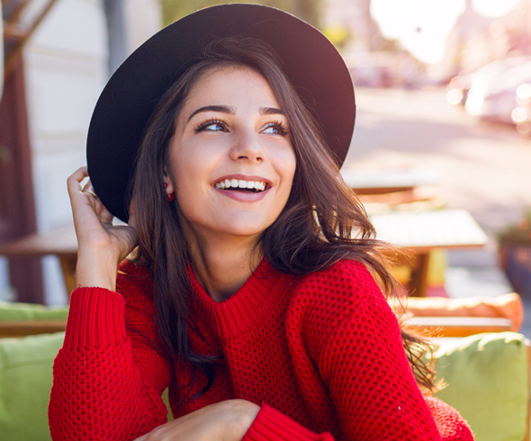 Young woman in red sweater and brown hat smiling outdoors during daytime.