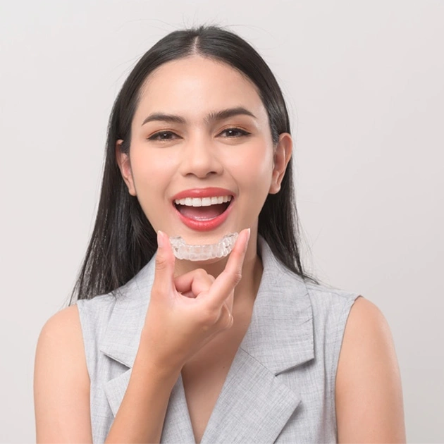 Woman in gray vest smiling brightly while applying lipstick against neutral background.